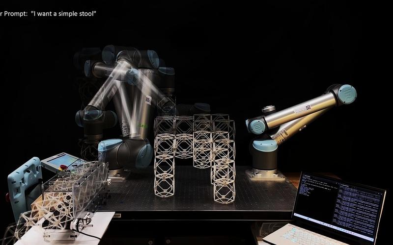 Two robotic arms assemble a geometric stool-like structure made of interlocking lattice components. A motion blur effect captures one arm’s movement as it places parts. A laptop and control panel sit nearby, and text in the corner reads “User Prompt: ‘I want a simple stool.’” The setup demonstrates real-time AI-driven robotic fabrication responding to natural language input.